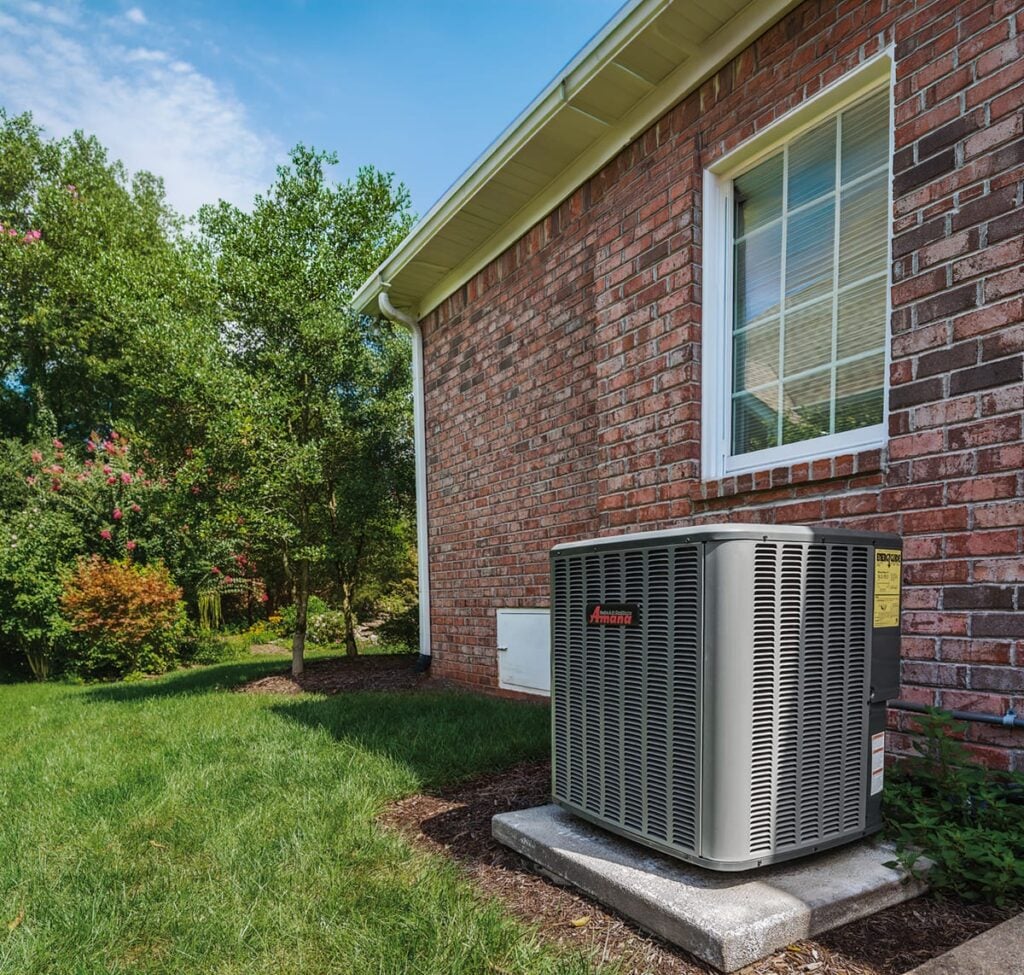 Amana air conditioning unit installed outside a brick house in a residential backyard.