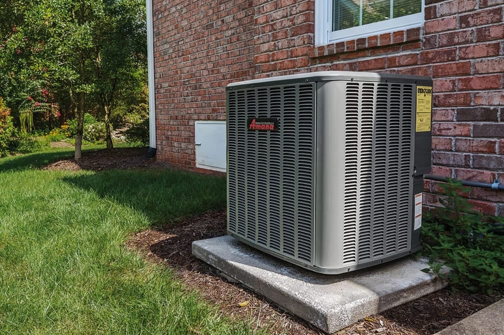 Amana air conditioning unit installed outside a brick house in a residential backyard.